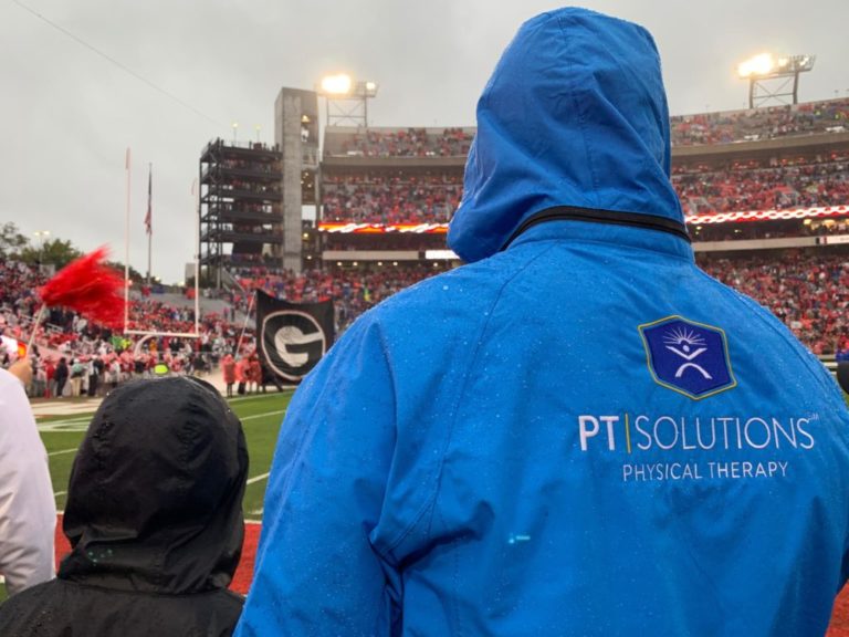 A Man With His Back Turned Wearing a Blue Jacket With the PT Solutions Logo in a Stadium